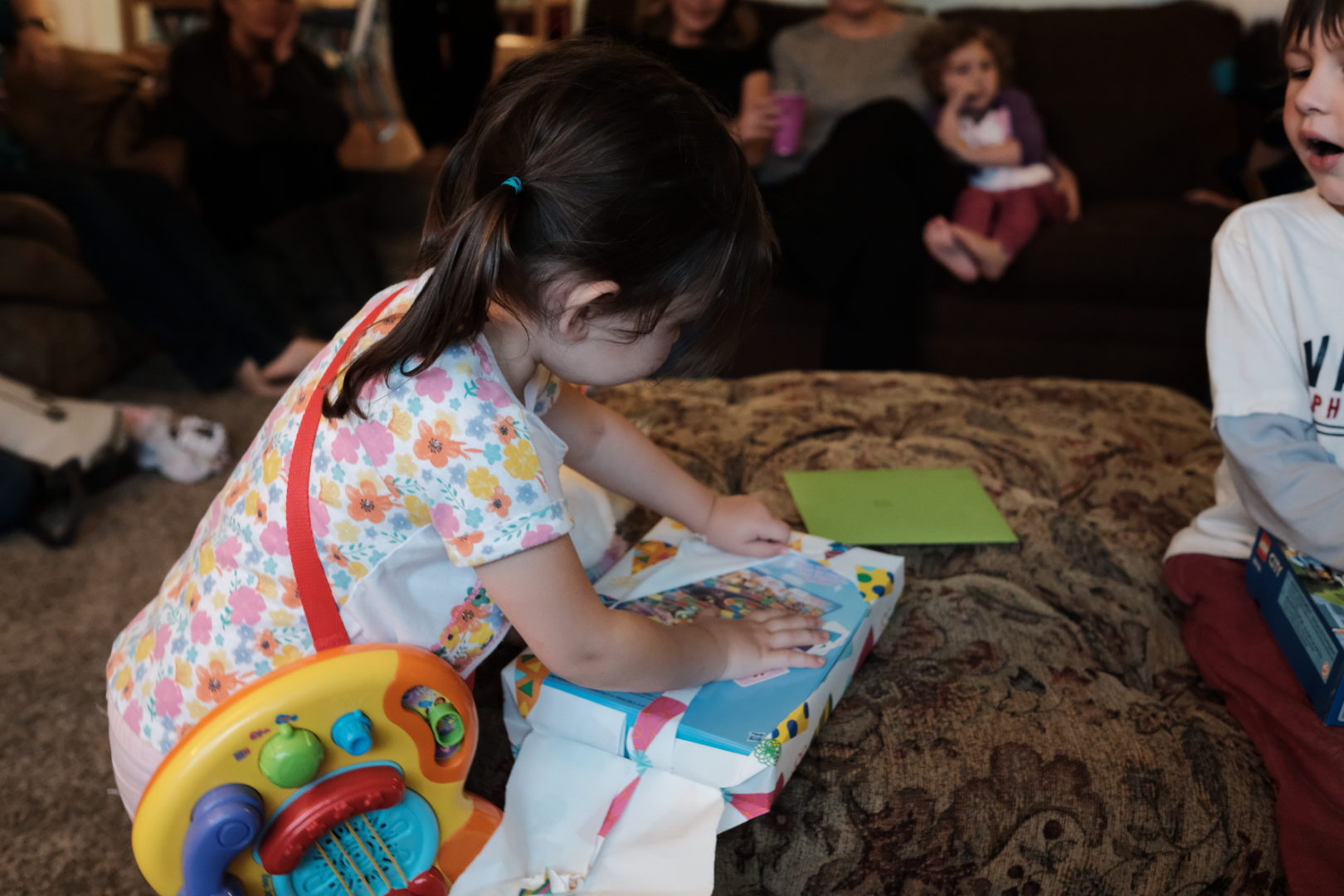 A little girl with a toy guitar around her shoulder opens a birthday present with Candyland.