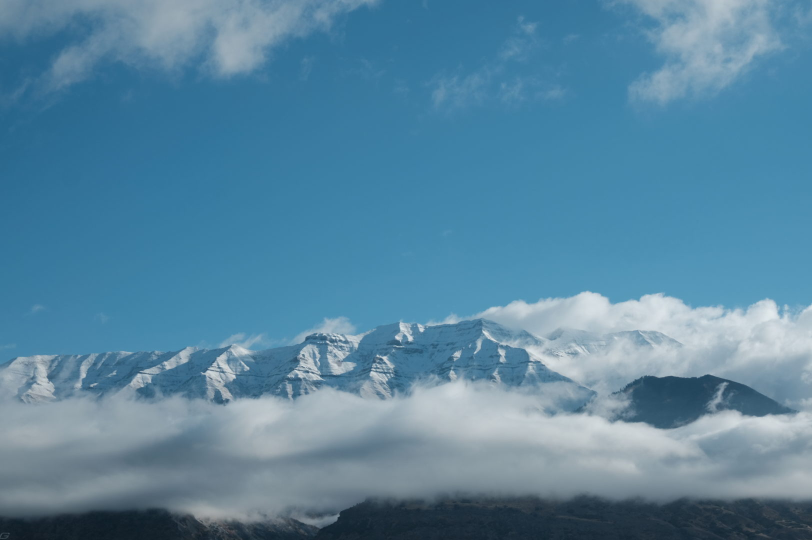 A light layer of snow covers the top of Mt Timpanogos as it's flanked by scattered fluffy clouds and a blue morning sky.