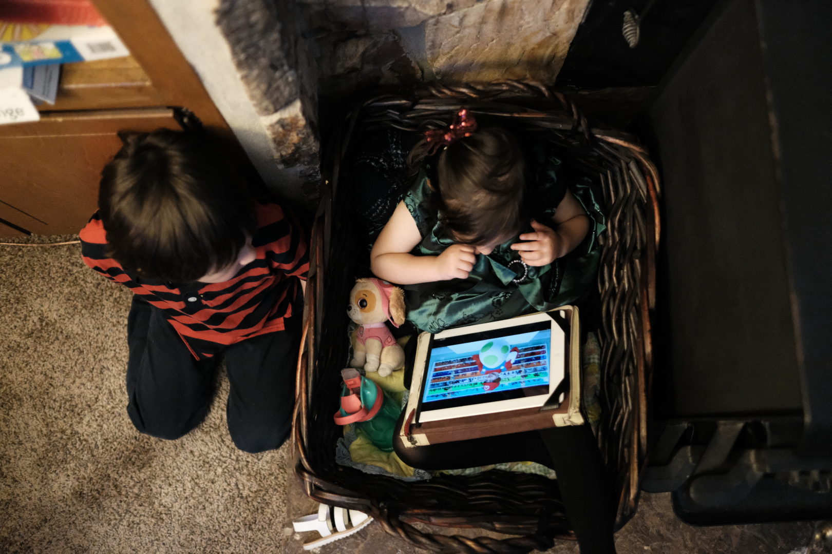 A little girls sits in a basket watching an iPad while her older brother sits next to her.